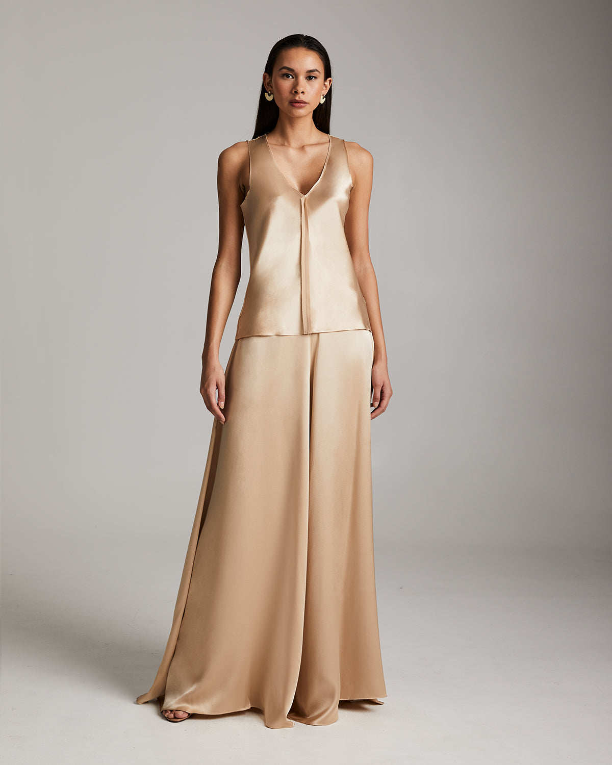 Woman wearing a beige satin jumpsuit against a plain background Silk palazzo pants in flowing charmeuse with dramatic wide leg silhouette, ethically made in NYC by VOZ