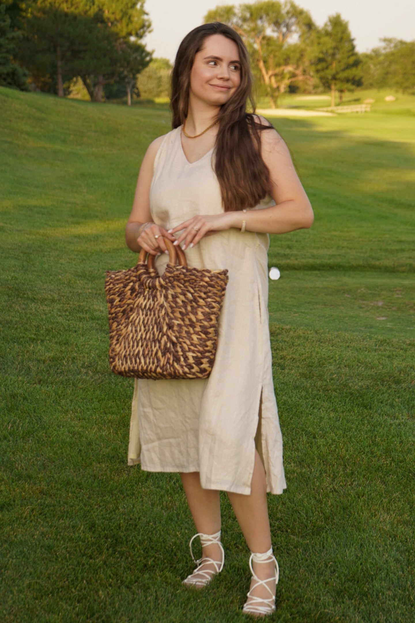 Woman in a light dress holding a woven bag on a grassy field