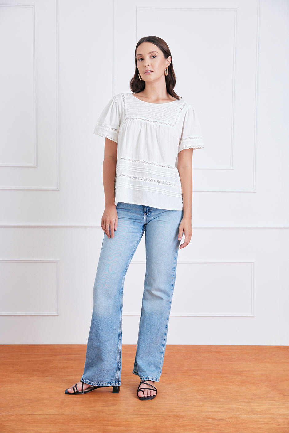Woman wearing a white blouse and light blue jeans standing against a white paneled wall. Dawn tee in off white sustainable cotton featuring romantic pin tuck and lace details with relaxed fit