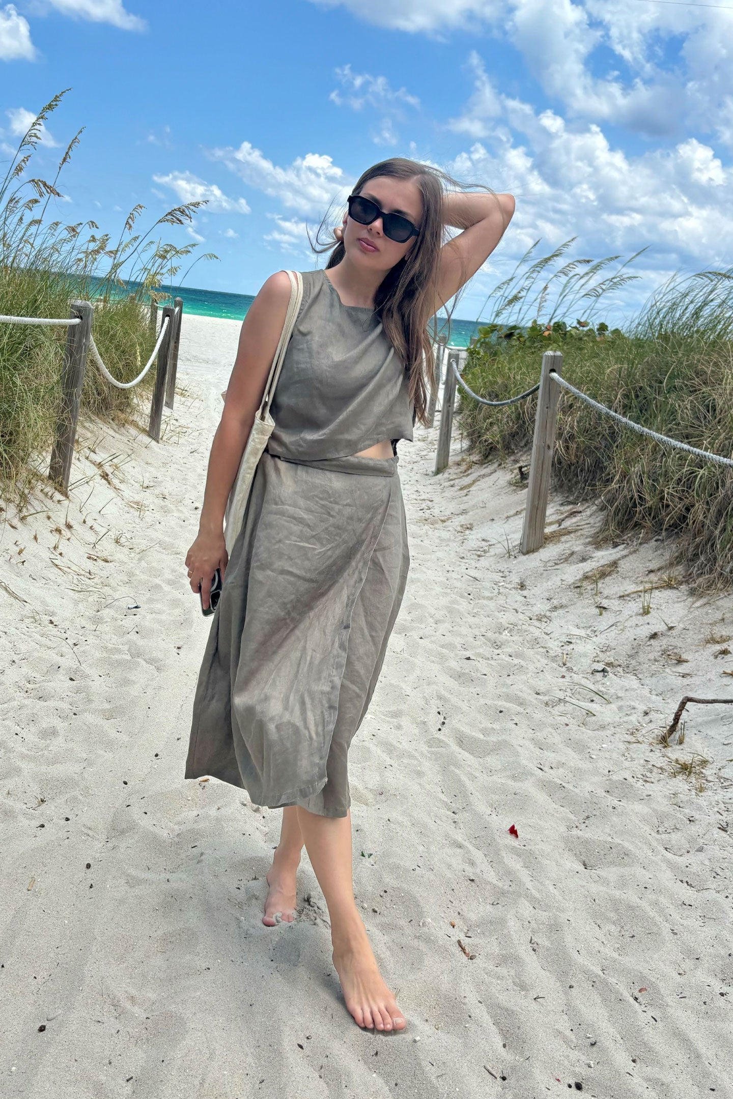 Woman in a gray dress standing on a sandy path with beach and ocean in the background