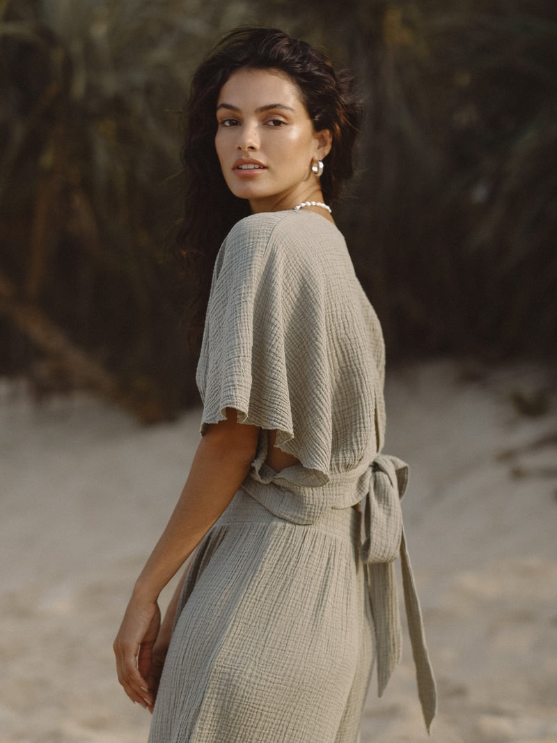 A A woman on a beach wearing a sustainable sage green dress with a tie-back, made from Turkish cotton by The Handloom, available at Elizabeth and Bern.