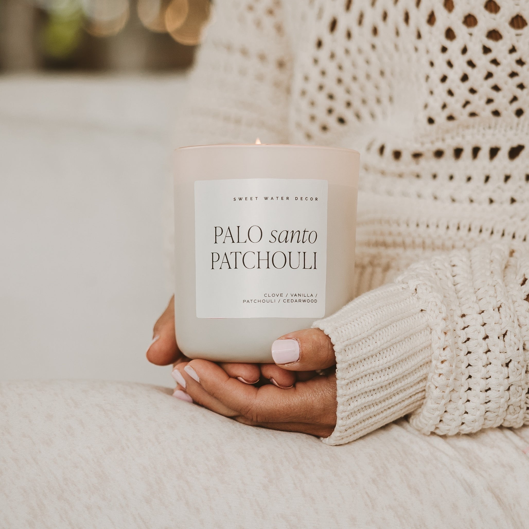 A woman in a cozy knit sweater holding a Palo Santo Patchouli scented soy candle from the Sweet Water Decor collection at Elizabeth and Bern.”