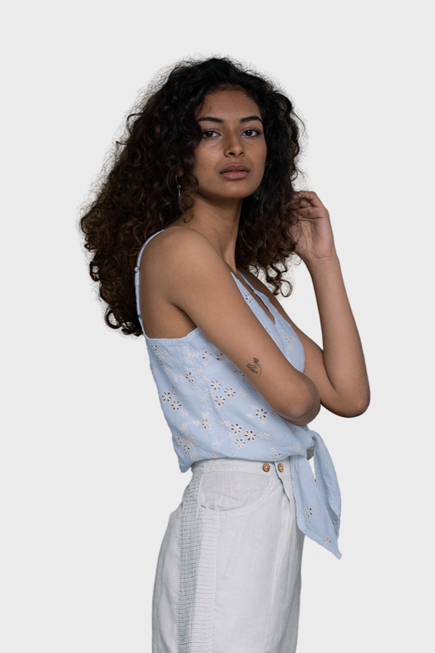 Woman wearing a light blue sleeveless top with floral patterns and white pants on a plain background. Pool Day Tan Lines camisole top in summer blue hemp with eyelet embroidery, tie-up front and adjustable straps