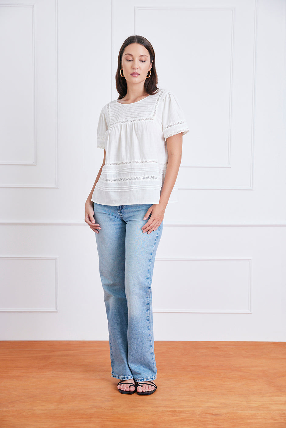 Woman wearing a white blouse and light blue jeans standing against a white paneled wall.