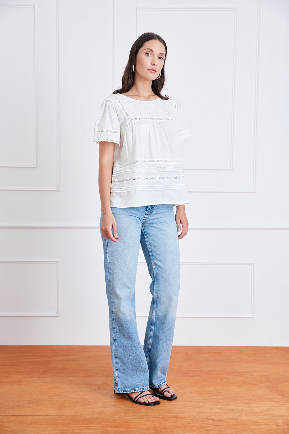 Woman wearing a white blouse and light blue jeans standing against a white paneled wall.