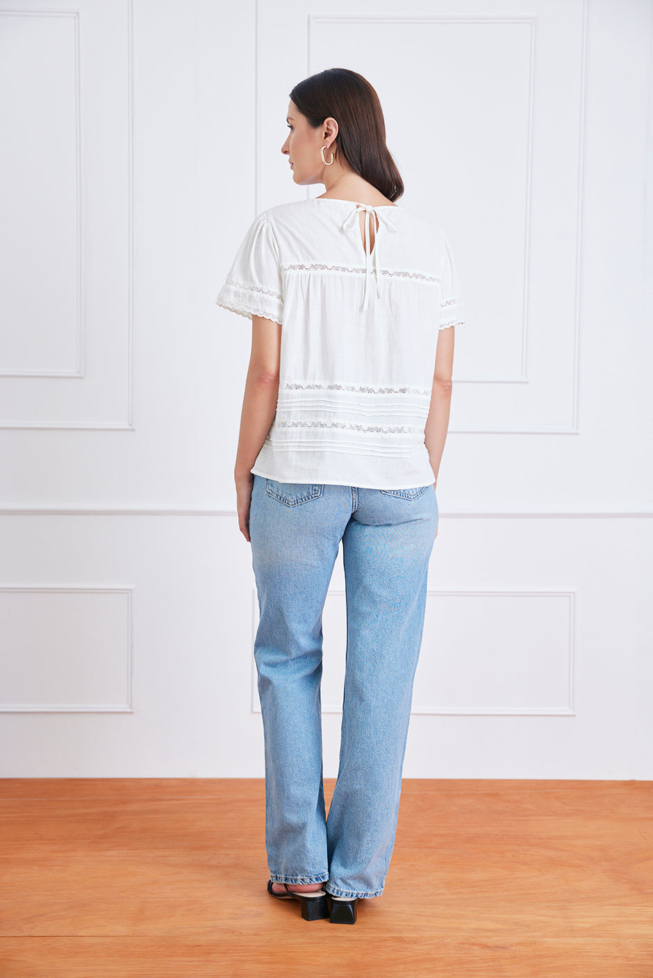 Woman wearing a white blouse and light blue jeans standing against a white wall.