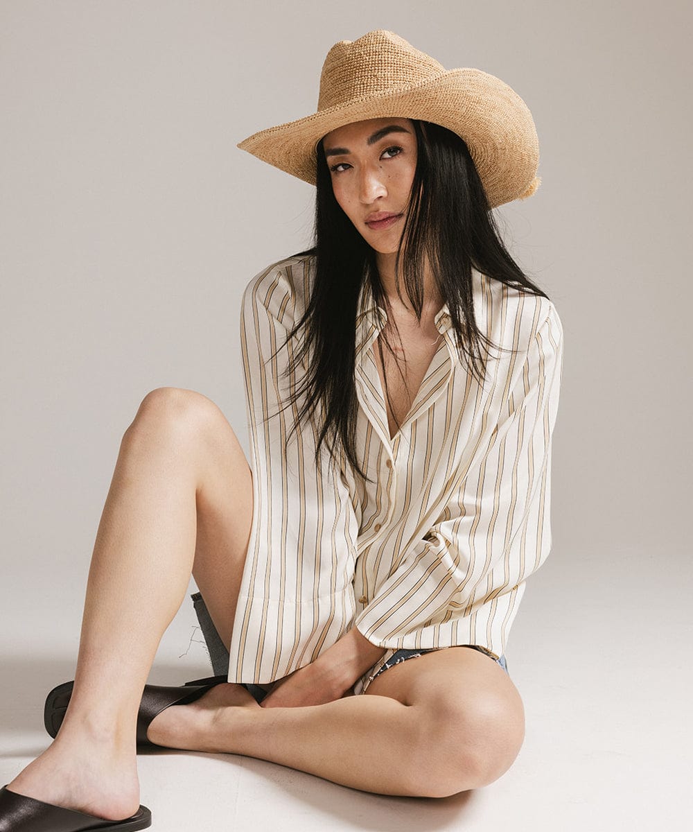 Woman wearing a straw hat and striped shirt sitting on the floor against a plain background